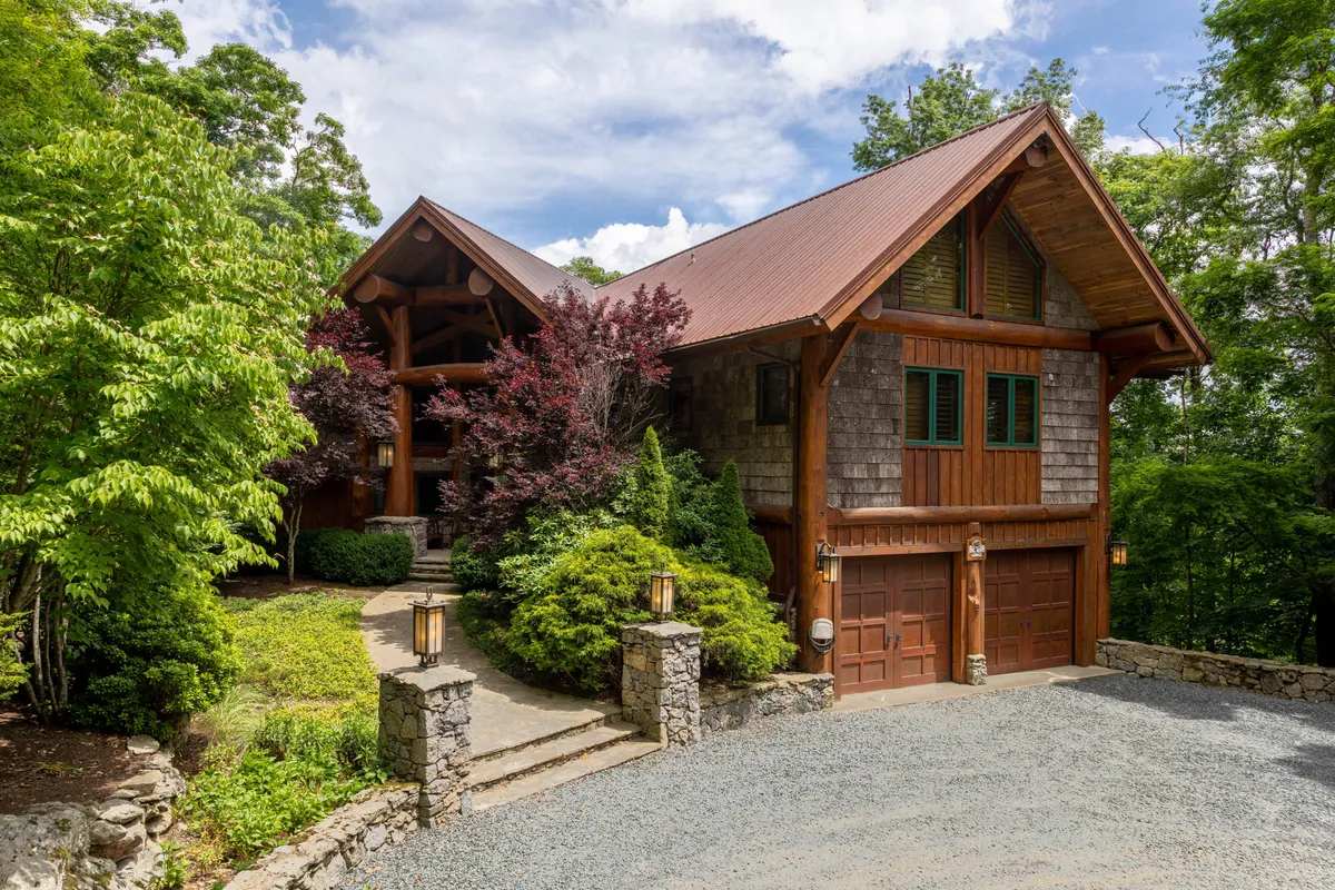 The hero image features the grand wooden entrance of a luxury mountain home, surrounded by lush greenery and a stone pathway leading invitingly to the front door. The setting is tranquil, showcasing the serene natural landscape and hinting at spacious outdoor living areas perfect for relaxation and gatherings.