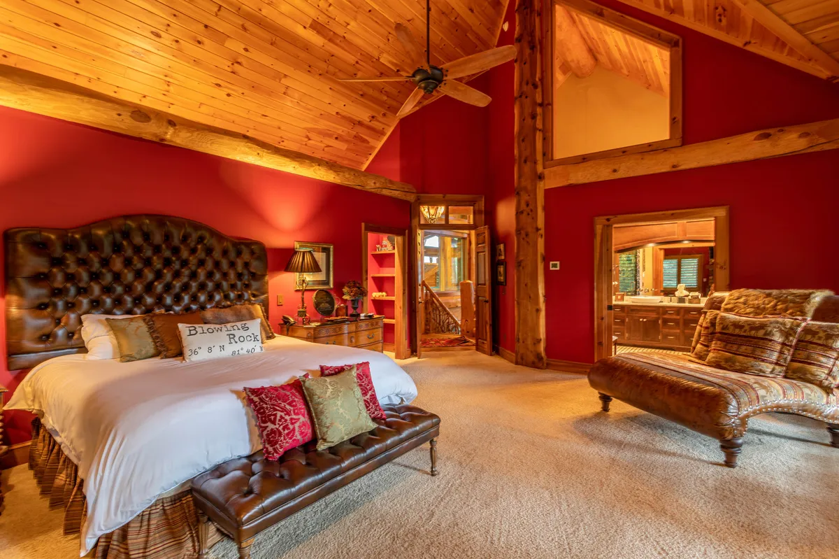 An inviting bedroom with a large tufted bed, ornate chaise lounge, exposed wooden ceiling beams, and red accent walls. The space exudes intimacy and warmth, perfect for relaxing mornings or quiet evenings.