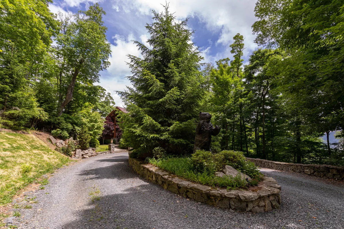 A peaceful outdoor scene featuring large evergreen trees, a charming stone wall, and a distinctive wooden bear statue set along a natural path. The lush landscaping and whimsical statue create a welcoming sense of adventure and discovery, perfect for families and children exploring the outdoors.