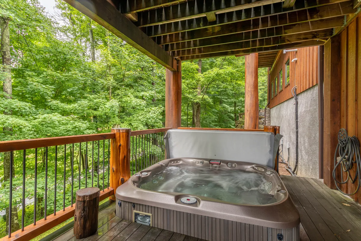 An exterior shot of a bubbling hot tub set on a wooden deck, surrounded by lush greenery. The scene is tranquil and inviting, suggesting the perfect spot for soothing relaxation after indoor games or a day outdoors. The wooden posts and deck maintain the cabin’s rustic vibe, while the open air hints at stargazing opportunities.