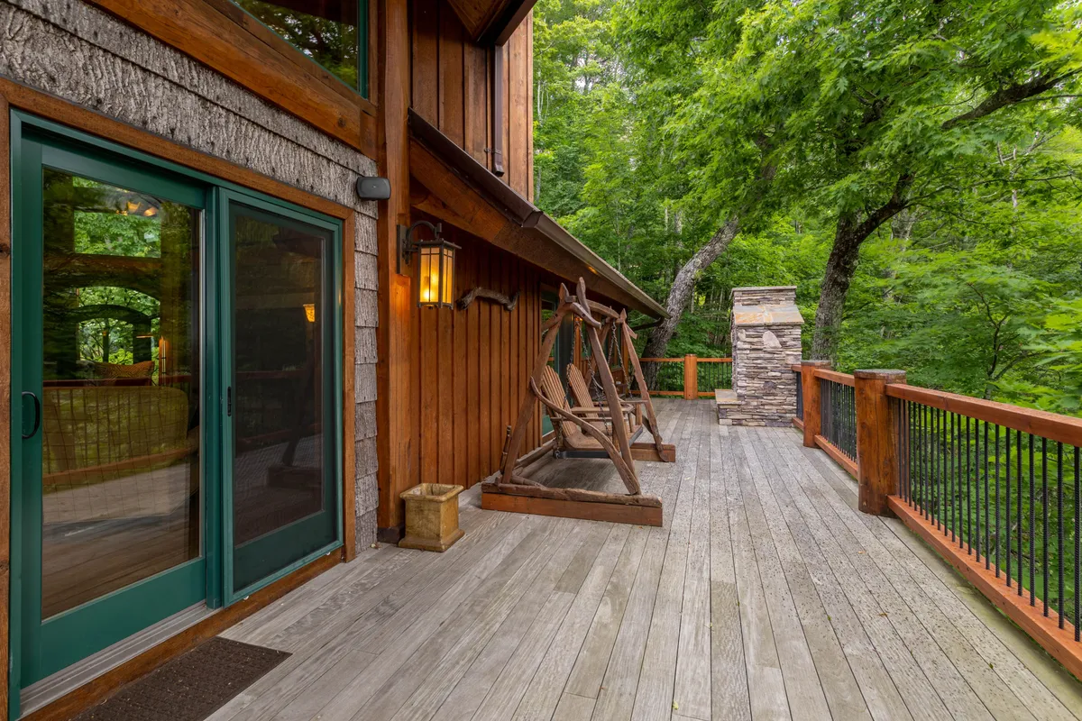 A tranquil view of a spacious wooden deck, featuring classic rocking chairs positioned to face out toward the trees and a stone feature integrated into the exterior wall. The setting exudes a sense of peacefulness, perfect for enjoying morning coffee or sunset relaxation while immersed in nature.