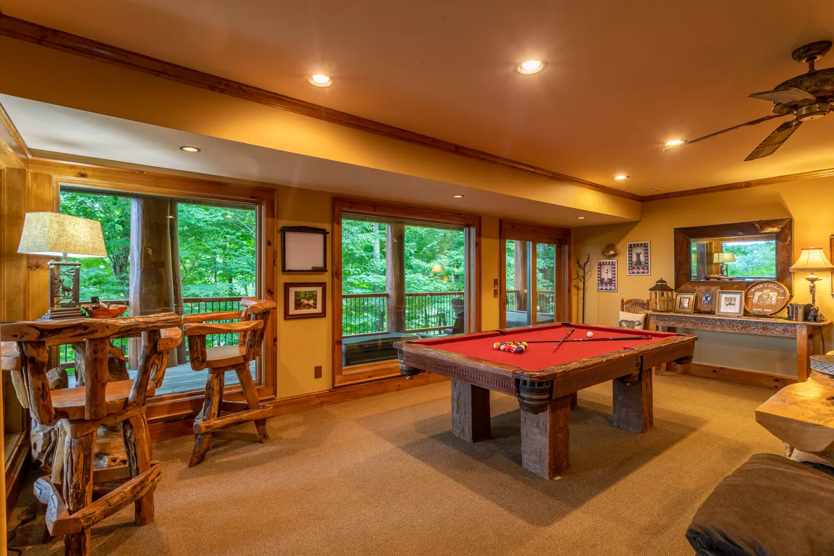 A cozy lower-level family room featuring a pool table as the main attraction, set beside large windows that fill the space with natural light. Wooden chairs, warm lamps, and decorative mountain-themed photographs contribute to the inviting, rustic cabin feel. The arrangement encourages gatherings, friendly pool games, and relaxed conversation, while the windows connect the indoors to the scenic outdoors.