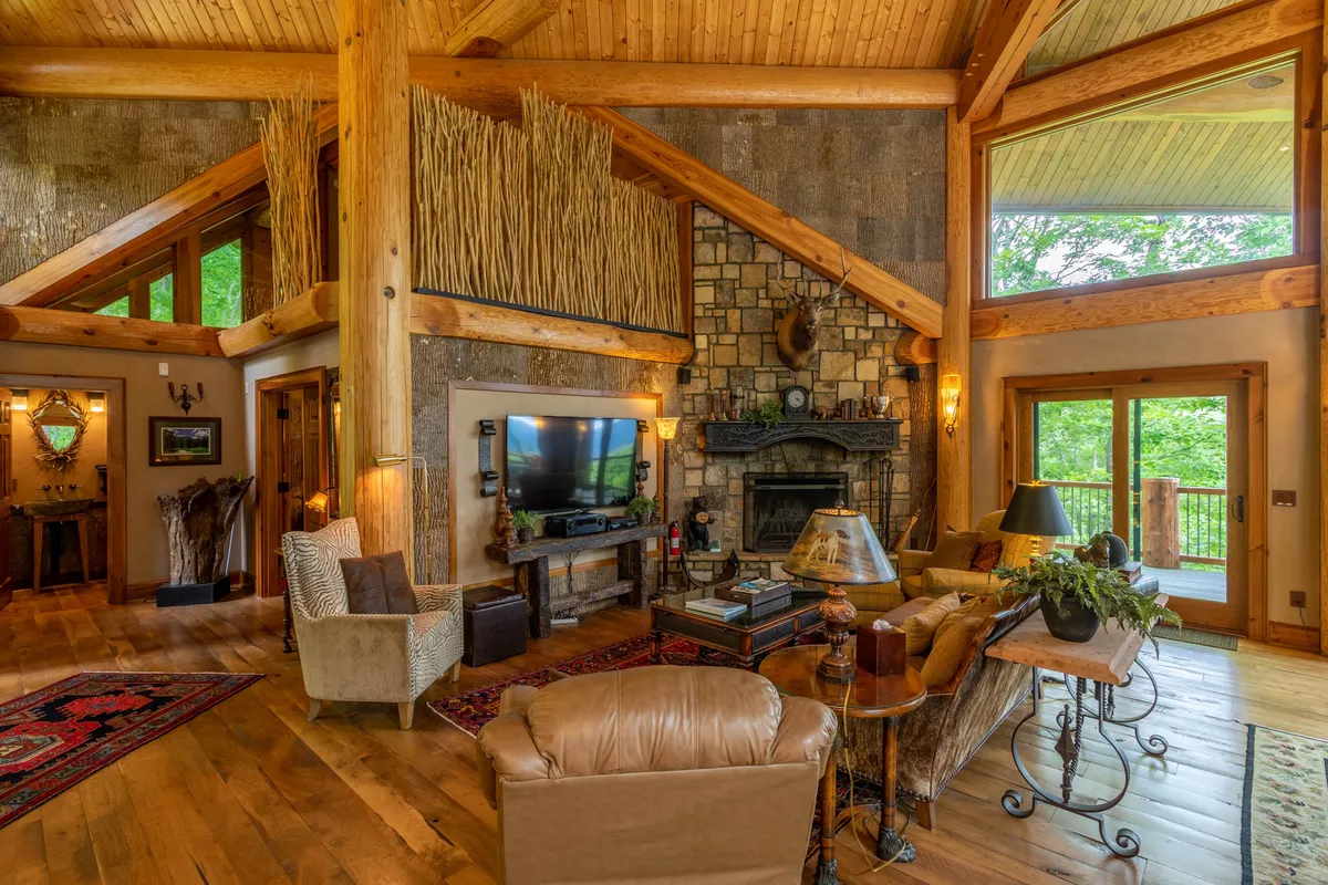 A spacious great room with a commanding stone fireplace, dramatic wooden beams across the cathedral ceiling, and expansive windows framing awe-inspiring mountain views. The space is furnished with rustic yet elegant furniture arranged to encourage conversation and relaxation, with the soft glow of the fireplace enhancing the inviting mood.