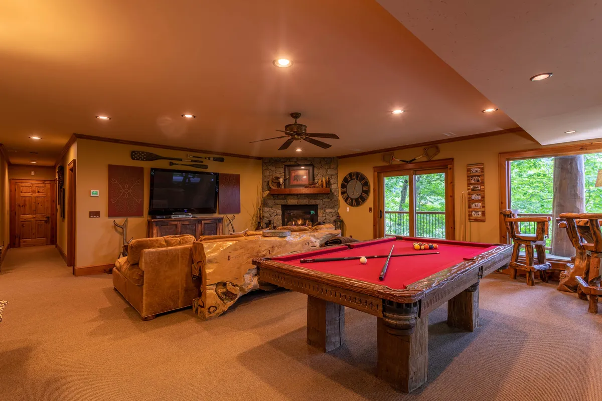 A recreation space that artfully combines a pool table and a stone fireplace, framed by log furnishings and large windows. The room's layout is ideal for both lively games and quiet moments by the fire, highlighting the versatility of the entertainment den. The wooden furniture and finishes maintain the authentic lodge style mentioned in the narrative.