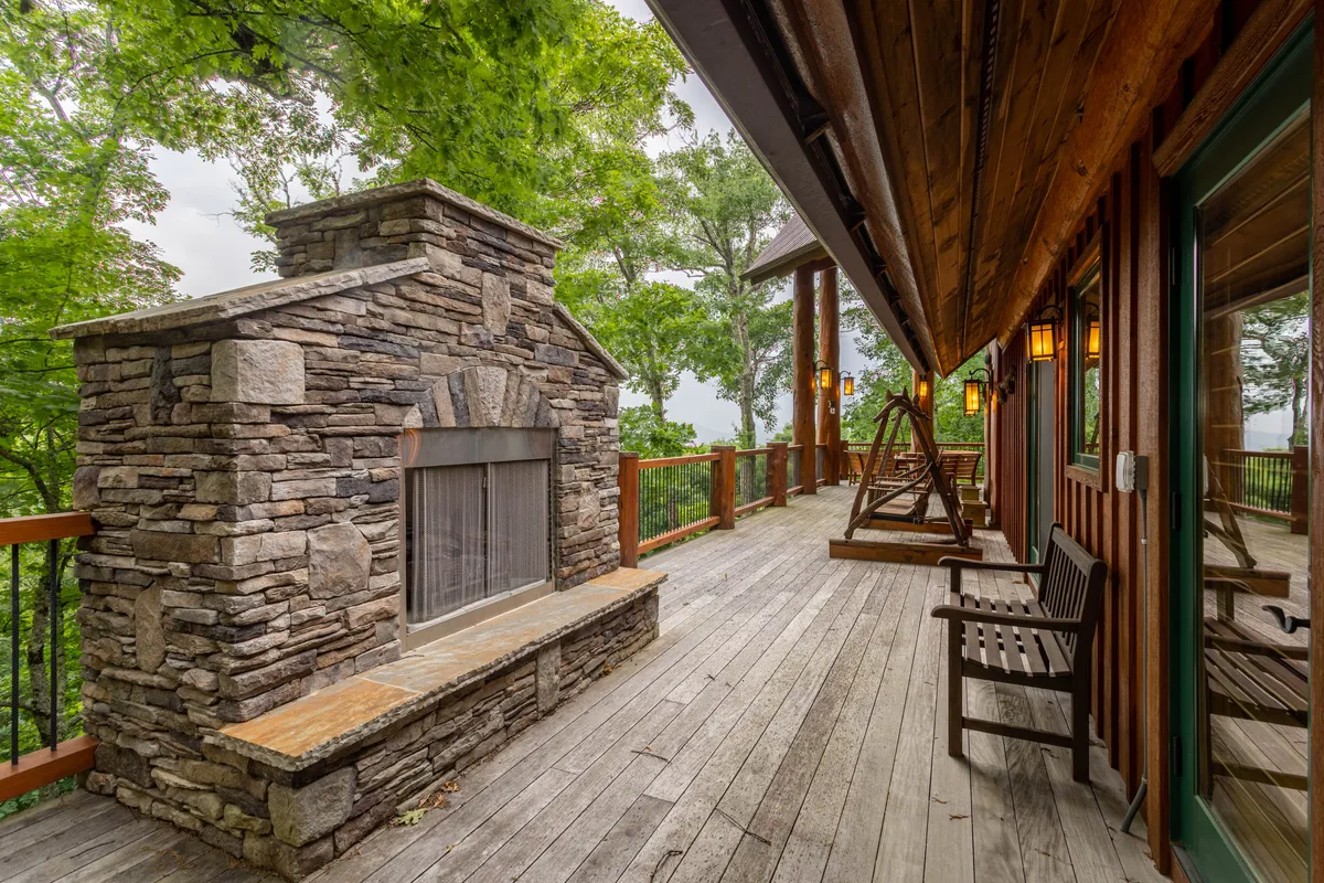 A spacious wooden deck featuring a large stone fireplace, a swinging bench, and surrounding trees. The seating area is positioned to take advantage of sunset views and the tranquility of the outdoors, creating a perfect backdrop for group gatherings or relaxed evenings.