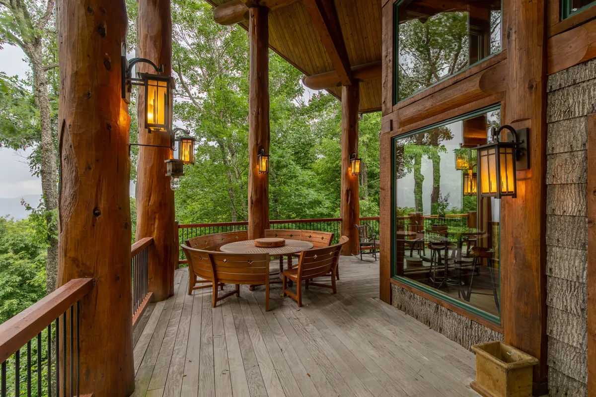 An inviting exterior dining area on a wooden deck, featuring a circular dining table surrounded by wooden pillars and lantern-style lighting. The scene is enhanced by the soft glow of sunset and lush greenery, suggesting a warm, communal space for al fresco meals.