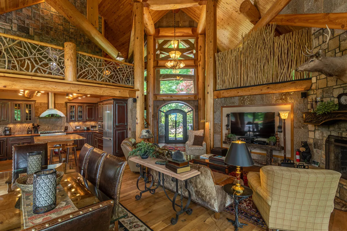 A welcoming open-plan kitchen and living area with a robust stone fireplace, exposed wooden beams, and a generous kitchen island. Decorative wall art and sunlight streaming through large windows emphasize the home's blend of rustic and contemporary design, while ample seating invites gatherings.