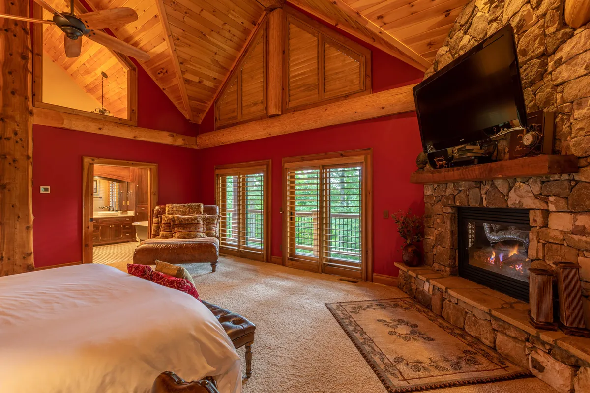 A warmly lit master suite with a plush king bed dressed in crisp white linens, a stone fireplace along one wall, and large windows allowing natural light to fill the space. Wooden beams lend a rustic mountain feel, while a plush chair near the fire hints at relaxation and luxury.