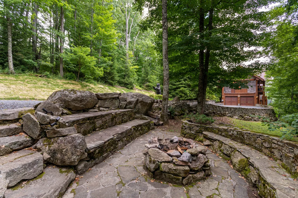 An inviting stone seating area encircles a fire pit among the trees, with a wooden house visible in the background. The setting is ideal for gathering, storytelling, and stargazing in a communal, nature-focused environment, reflecting the social and adventurous spirit of Eagles Nest.