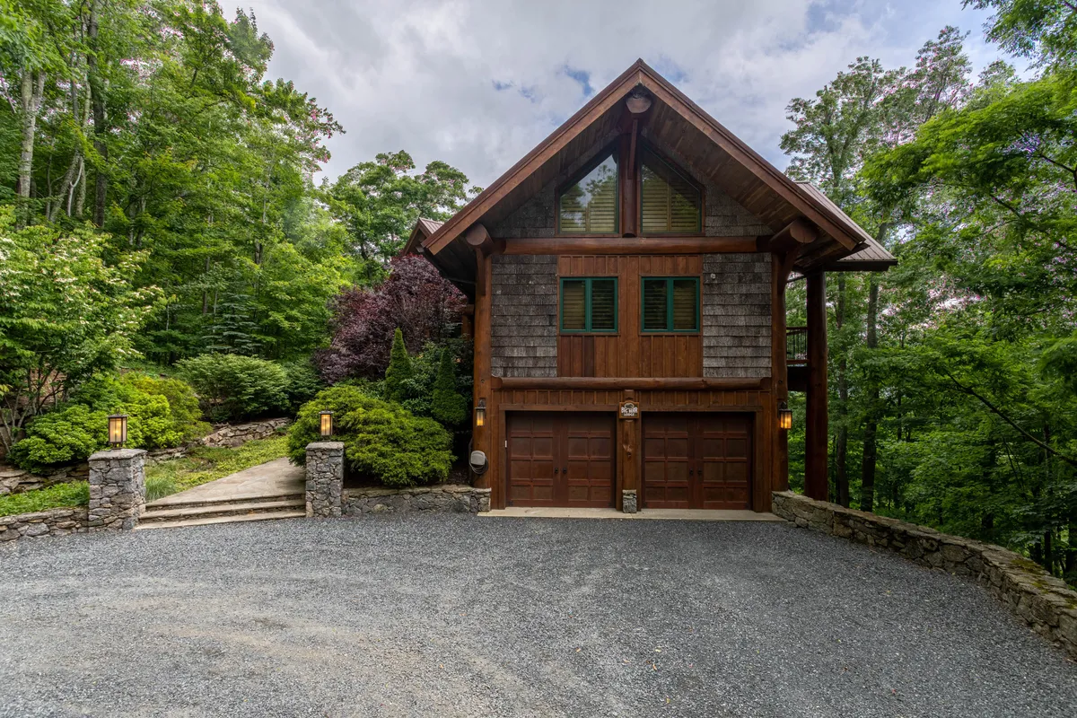 The image captures a cozy mountain home exterior with a wooden facade and stone pillars surrounded by lush greenery. The setting is serene and inviting, hinting at community trails, fire pit gatherings, and pet-friendly exploration.